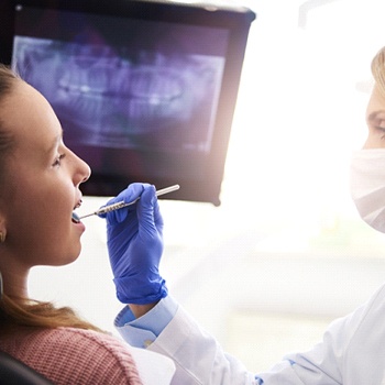 A dentist examining a female patient’s smile while her digital x-rays are displayed on a nearby screen