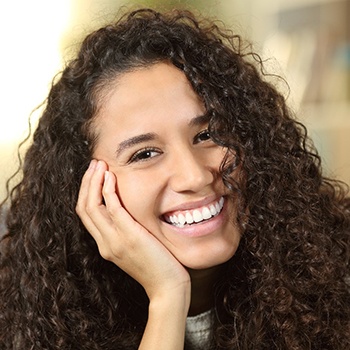 A teenage girl with long, dark, curly hair propping her face on her hand and smiling